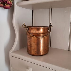 May include: A cylindrical copper bucket with a polished metallic finish and a weathered patina. The vessel features a rounded rim, a horizontal band detail around the upper section, and a thin, arched brass handle attached to two side loops.