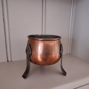 May include: A small, antique copper cauldron with three metal legs. The cauldron has a rounded body and a dark interior. The legs are attached to the cauldron with rivets. The copper has a warm, reddish-brown colour.