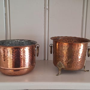 May include: Two hammered copper vessels with circular side handles. The left vessel has a smooth base, while the right vessel features a scalloped rim and stands on three decorative brass feet. Both containers have a metallic, reflective finish.