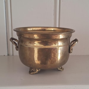 May include: A polished brass bowl with a rounded body, a wide rim, and two small side handles. The vessel is supported by three short, decorative feet. The metallic surface reflects light, showing a warm, golden-yellow tone throughout the piece.