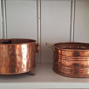 May include: Two round copper vessels with metallic finishes. The left vessel features a hammered texture, side ring handles, and three small feet. The right vessel has horizontal ridges and two side handles. Both items are open-topped containers.