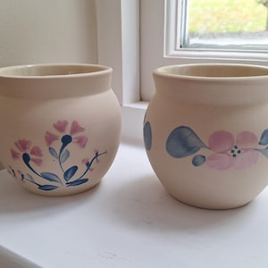 May include: Two beige ceramic pots with floral patterns. One pot has pink and blue flowers, the other a similar design. The pots are round with a wide opening, ideal for plants or decorative purposes.