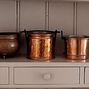 May include: Three copper vessels arranged in a row. The left is a rounded pot with three small feet. The centre is a cylindrical hammered container with a swing handle. The right is a straight-sided pot with two side handles and a base rim.