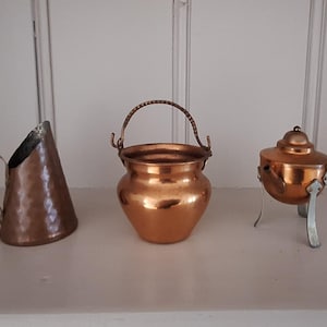 May include: A set of three copper items including a hammered conical jug with a side handle, a small round pot with a swing handle, and a lidded copper pot with a long metal handle and three legs, all displayed in a horizontal row.