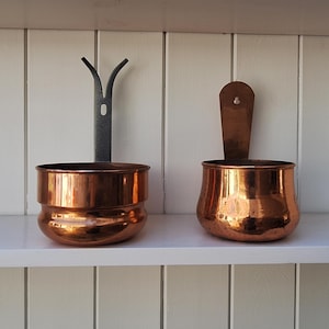 May include: Two wall-mounted copper containers. The left container features a dark metal bracket with a Y-shaped top. The right container has a flat, rounded copper backplate with a small hole for hanging. Both items have a polished finish.