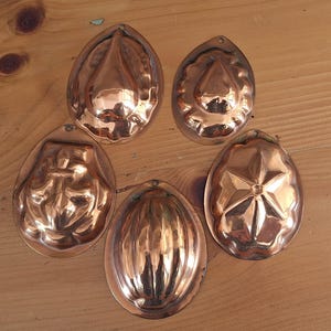 May include: A set of five copper-coloured metal moulds with various embossed designs including a teardrop, a star, and vertical fluting. Each mould features a small hole at the top edge for hanging, with a polished, reflective surface finish.