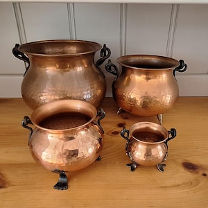 May include: A set of four graduated copper pots with a hammered finish, each featuring dark metal handles and three small feet. The pots are arranged in a group, showcasing their rounded bodies and open tops, reflecting a metallic sheen.