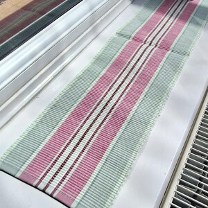 May include: A rectangular table runner featuring a woven striped pattern in shades of sage green, dusty rose pink, and thin dark burgundy lines. The fabric has a textured, ribbed appearance with finished edges, displaying a consistent vertical stripe design.