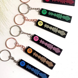 May include: A collection of black keychains with a Spotify code design. Each keychain features a different color scheme, including red, green, yellow, pink, orange, and blue. The keychains have silver and rose gold key rings.