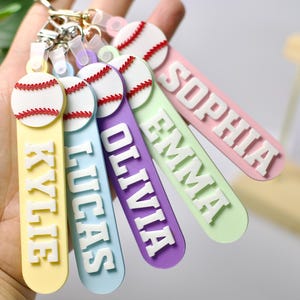 May include: A set of five silicone keychains, each featuring a white baseball with red stitching at the top and a vertical rectangular tag below. The tags are yellow, light blue, purple, light green, and pink, with white raised letters spelling KYLIE, LUCAS, OLIVIA, EMMA, and SOPHIA.