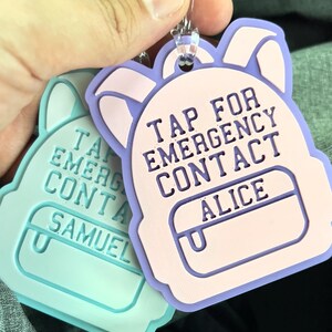 May include: Two backpack-shaped luggage tags. One is light blue with the name "SAMUEL", the other is pink and purple with the name "ALICE". Both tags have the text "TAP FOR EMERGENCY CONTACT".