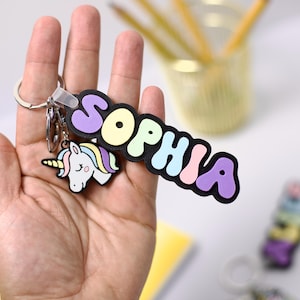 May include: A keychain with the name "SOPHIA" in colorful bubble letters. A small unicorn charm with a rainbow mane and tail hangs below the name. The keychain is held in a hand.