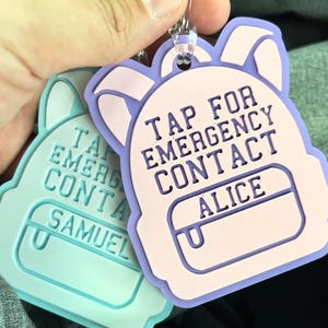 May include: Two backpack-shaped luggage tags. One is light blue with the name "SAMUEL", the other is pink and purple with the name "ALICE". Both tags have the text "TAP FOR EMERGENCY CONTACT".