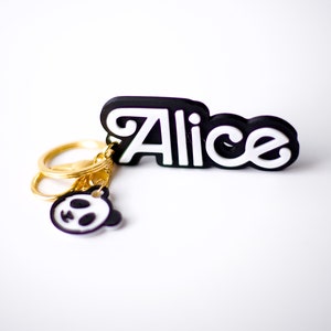 May include: Black and white keychain with the name "Alice" in white letters on a black background. The keychain has a gold key ring with a small black and white panda charm.
