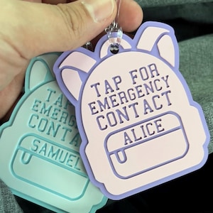 May include: Two backpack-shaped tags with clear plastic loops. One is light blue with the text "TAP FOR EMERGENCY CONTACT SAMUEL". The other is lavender and pink with the text "TAP FOR EMERGENCY CONTACT ALICE".