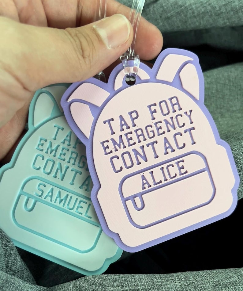 May include: Two backpack-shaped tags with clear plastic loops. One is light blue with the text "TAP FOR EMERGENCY CONTACT SAMUEL". The other is lavender and pink with the text "TAP FOR EMERGENCY CONTACT ALICE".