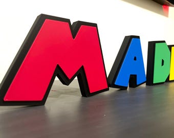 May include: A set of colorful, 3D letters spelling out the word "MAD" with black outlines. The letters are made of foam and are standing on a gray surface.