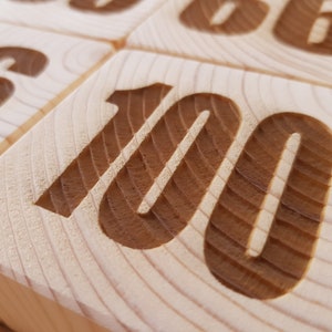 Puede incluir: Un bloque de madera con el número "100" tallado en él. El número está en una fuente marrón y el bloque es de color madera claro.