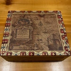 May include: A wooden box with a decorative lid featuring a vintage map of Europa. The map is surrounded by a gold and red border with intricate designs. The lid also includes the text "EUROPA" and "Here Beauty".
