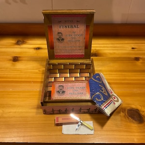 May include: A wooden box with a hinged lid, open to reveal a brick pattern interior. Inside the box is a small card with a photo of John Henry "Doc" Holliday and a blue and white bandana. A small pocket knife and a pack of matches are on the table in front of the box.