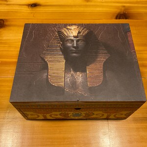 May include: A wooden box with a decorative design featuring a pharaoh's head with a gold headdress. The box has a dark brown background with gold accents.