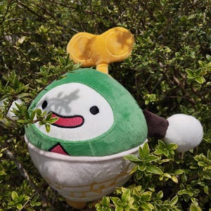 May include: A plush toy with a green, white, and yellow design, featuring a smiling face and a wind-up key on top. The toy is nestled among green foliage, creating a playful and whimsical scene.