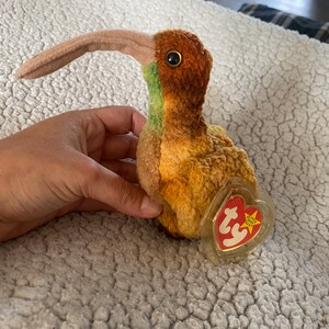Könnte beinhalten: Ein brauner und grüner Plüsch-Kiwi mit einem langen orangefarbenen Schnabel. Der Vogel hält ein rotes und weißes Ty Beanie Baby-Etikett mit dem Wort "Ty" in weiß.