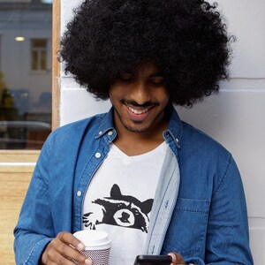 May include: A man with an afro hairstyle is wearing a blue denim shirt over a white t-shirt with a black raccoon graphic. He is holding a disposable coffee cup and a black smartphone.