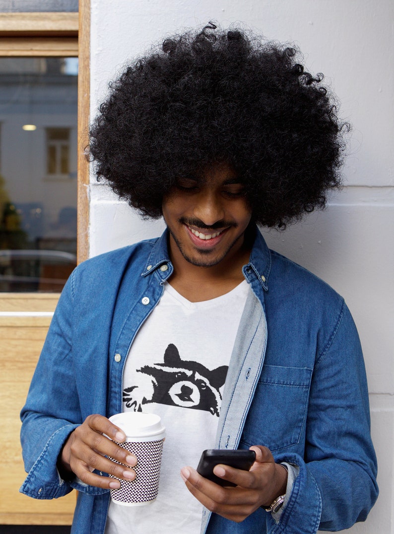 May include: A man with an afro hairstyle is wearing a blue denim shirt over a white t-shirt with a black raccoon graphic. He is holding a disposable coffee cup and a black smartphone.