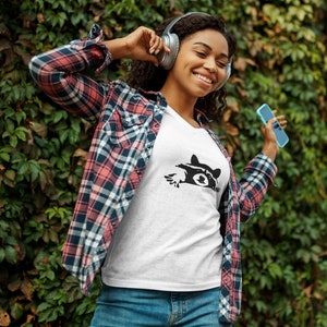 May include: A woman wearing a white t-shirt with a black raccoon graphic, a red and black plaid shirt, blue jeans, and headphones. She is holding a blue smartphone and smiling.