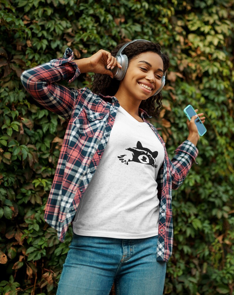 May include: A woman wearing a white t-shirt with a black raccoon graphic, a red and black plaid shirt, blue jeans, and headphones. She is holding a blue smartphone and smiling.