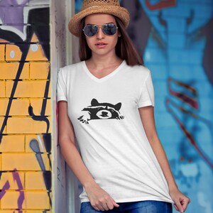 May include: A white V-neck t-shirt with a black graphic of a raccoon peeking out from behind a wall. The raccoon has a black nose and eyes.