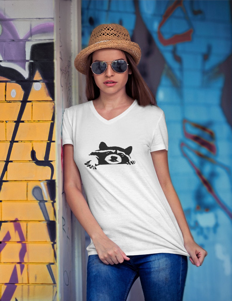 May include: A white V-neck t-shirt with a black graphic of a raccoon peeking out from behind a wall. The raccoon has a black nose and eyes.