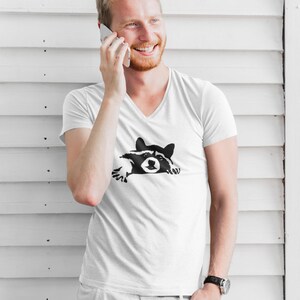 May include: A white V-neck t-shirt with a black and white graphic of a raccoon peeking out from behind a wall.