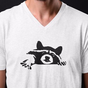 May include: A white V-neck t-shirt with a black silhouette of a raccoon peeking over the edge of the shirt.