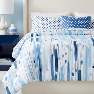 May include: A bed featuring a white and blue watercolor-style striped and dotted comforter with matching polka dot pillows. Beside the bed sits a dark blue nightstand with a white lamp, a small vase, and a large green potted plant nearby.