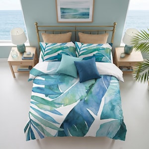 May include: A bedding set featuring a watercolor print of large tropical palm leaves in shades of teal, blue, and green on a white background. The set includes a duvet cover, two matching pillowcases, one solid light blue pillow, and one solid dark blue pillow.