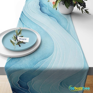 May include: A blue and white watercolor-style table runner with a wavy pattern. A light blue plate with a white rim and a place card that reads "MARY" is on the table. A vase of flowers is in the background.