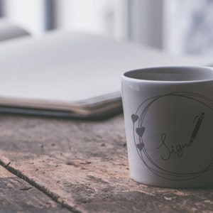 May include: White ceramic mug with a black and gray design. The design features a circle with a pencil and the word "Sign" written in cursive. There are four small hearts around the circle.