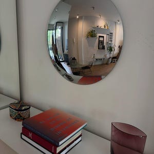 May include: A circular convex wall mirror reflecting a room interior. Below the mirror, a stack of books titled El Color en el Diseño de Interiores, a small patterned box, a small dog figurine, and a translucent purple glass vase sit on a white surface.