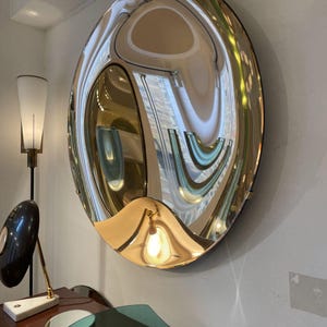 May include: A large, circular, convex wall mirror with a polished gold-toned frame. The reflective surface is curved outward, creating a distorted, wide-angle view of the surrounding room, including reflections of light fixtures and architectural details.