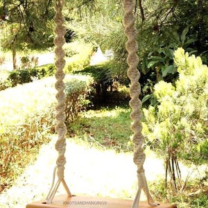 May include: A wooden swing seat with natural-colored rope hanging from a tree branch. The swing seat is made of wood and has a natural finish. The rope is knotted and has a natural color. The swing is hanging in a garden setting.