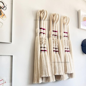 May include: A set of five off-white macrame wall hangings with looped tops and long, fringed bottoms. Each piece features burgundy and cream-colored bands and decorative knots. The text "KNOTS&HANGINGS" is visible on the wall.