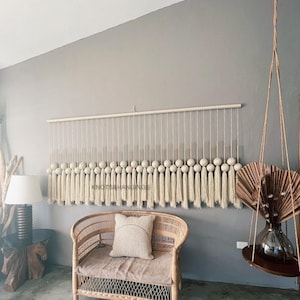 May include: A beige macrame wall hanging with a wooden dowel and tassels. The hanging features a series of round, beige balls. The text "Knots & Hangings" is visible in the image.