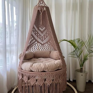 以下が含まれることがあります： 淡いローズカラーのハンギングマクラメチェア。丸いクッションシートと2つのボルスター枕、フリンジエッジが特徴です。キャノピーは幾何学模様で複雑に織られています。