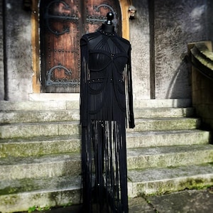 Può includere: Abito nero in macramè su manichino. L'abito presenta un collo alto, maniche lunghe e dettagli con frange. Il manichino è esposto su gradini di pietra e una porta di legno.