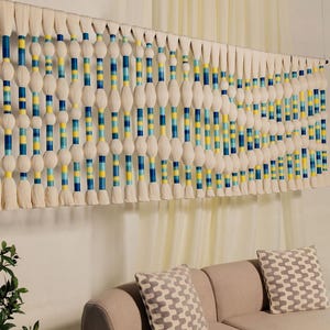 May include: A large, handmade wall hanging with a repeating pattern of white, blue, and yellow yarn. The design features alternating sections of solid color and textured white yarn, creating a visually appealing, modern aesthetic. The wall hanging is displayed above a beige sofa.