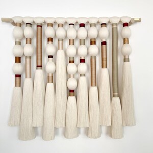 May include: A wall hanging featuring eleven vertical strands of cream-colored yarn suspended from a light wood rod. The strands consist of alternating wrapped spheres, smooth cylindrical sections, and long tassels in cream, tan, and deep red.