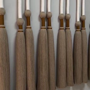 May include: A series of decorative wall hangings with a neutral color palette. Each piece features a curved, white-wrapped top, a wooden sphere, and a long, textured tassel in shades of beige and brown. The wall is painted a light gray.