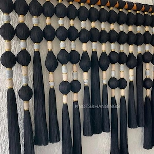 May include: Black and white wall hanging with tassels and beads. The wall hanging is made of yarn and has a wooden dowel at the top. The text "Knots & Hangings" is visible at the bottom of the image.